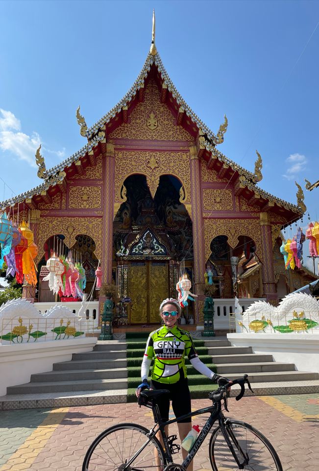 A photo of Elizabeth standing with her bike in front of a red house of worship near Chiang Mai