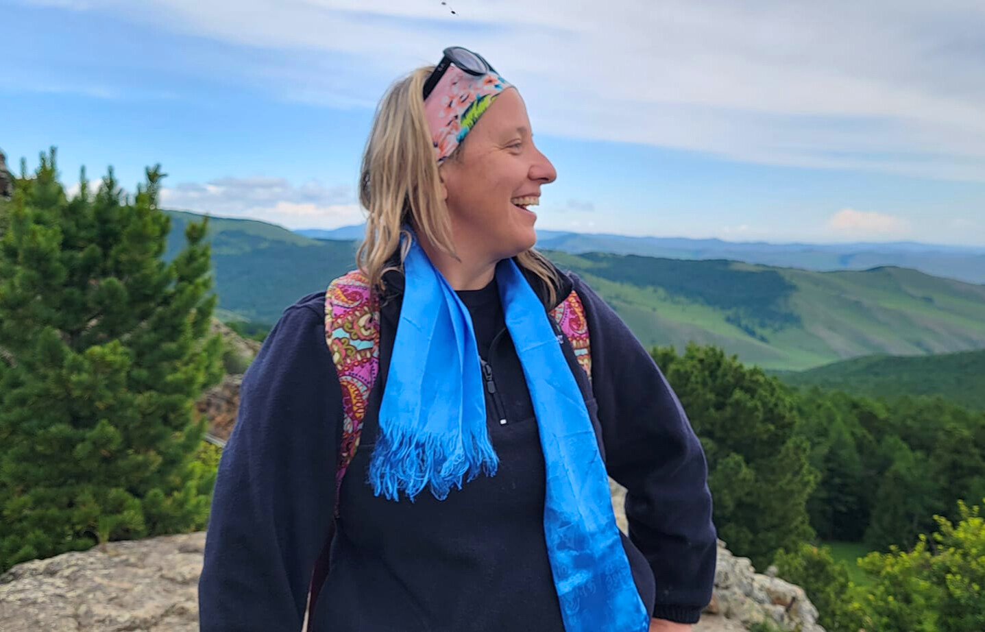 Tracy stands on a mountaintop with a beautiful mountainous landscape in the background.